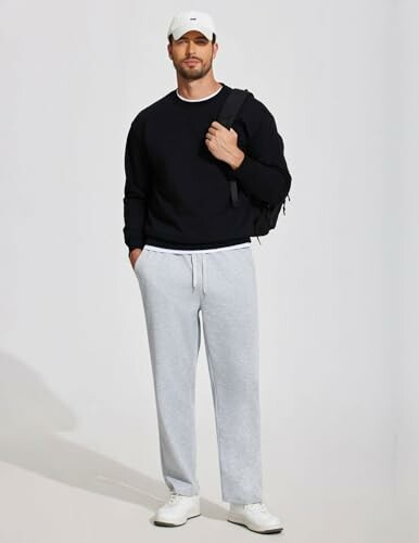 Man in black sweater, gray sweatpants, and white cap with backpack.