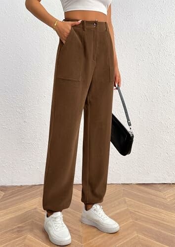 Woman wearing brown pants, white crop top, and white sneakers holding a black handbag.