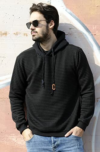 Man wearing a black hoodie and sunglasses standing against a wall.