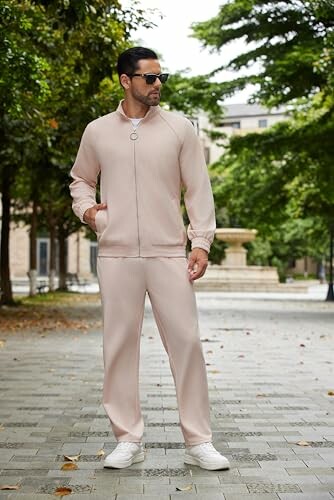 Man in beige tracksuit and sunglasses standing on a paved path with trees.